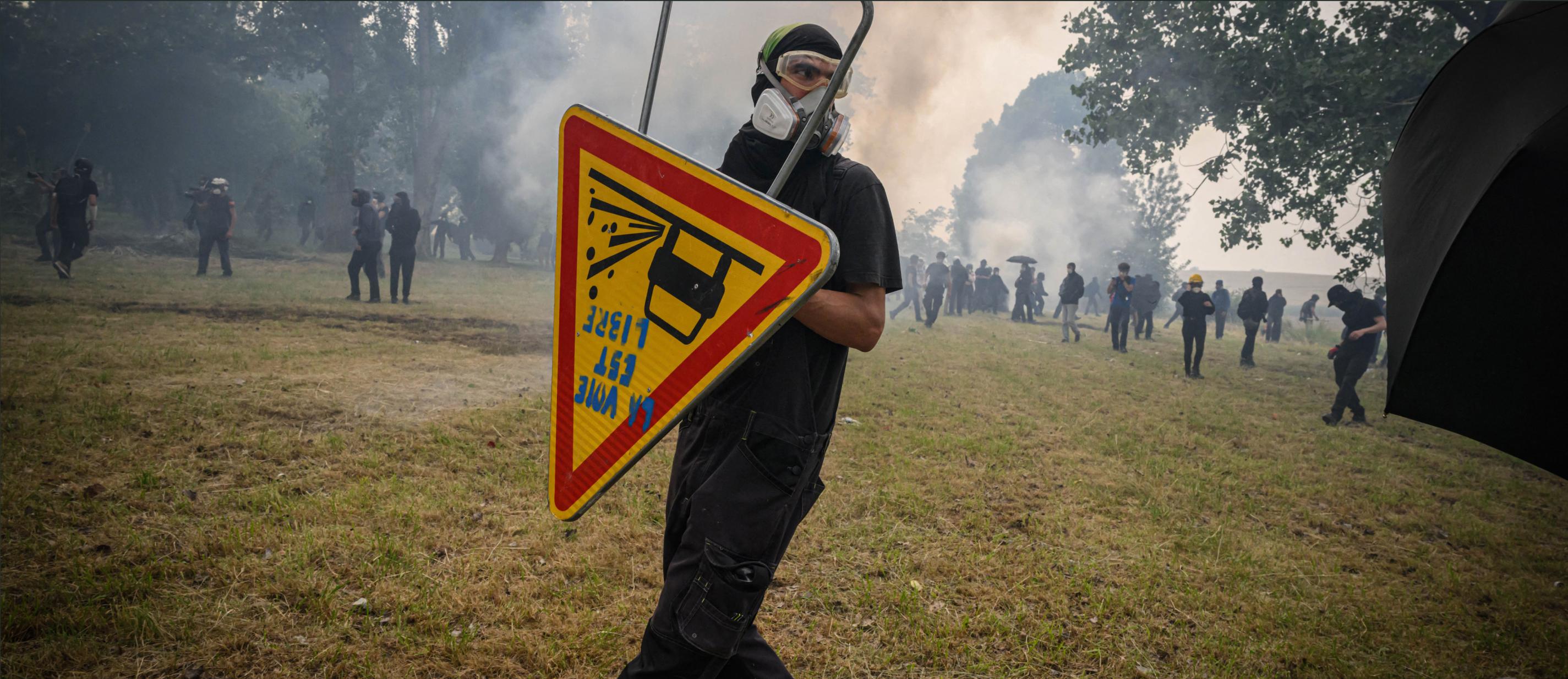 Un manifestant avec masque à gaz et lunettes de protection avec en arrière plan de la fumée et des dizaines de manifestants dans un champ