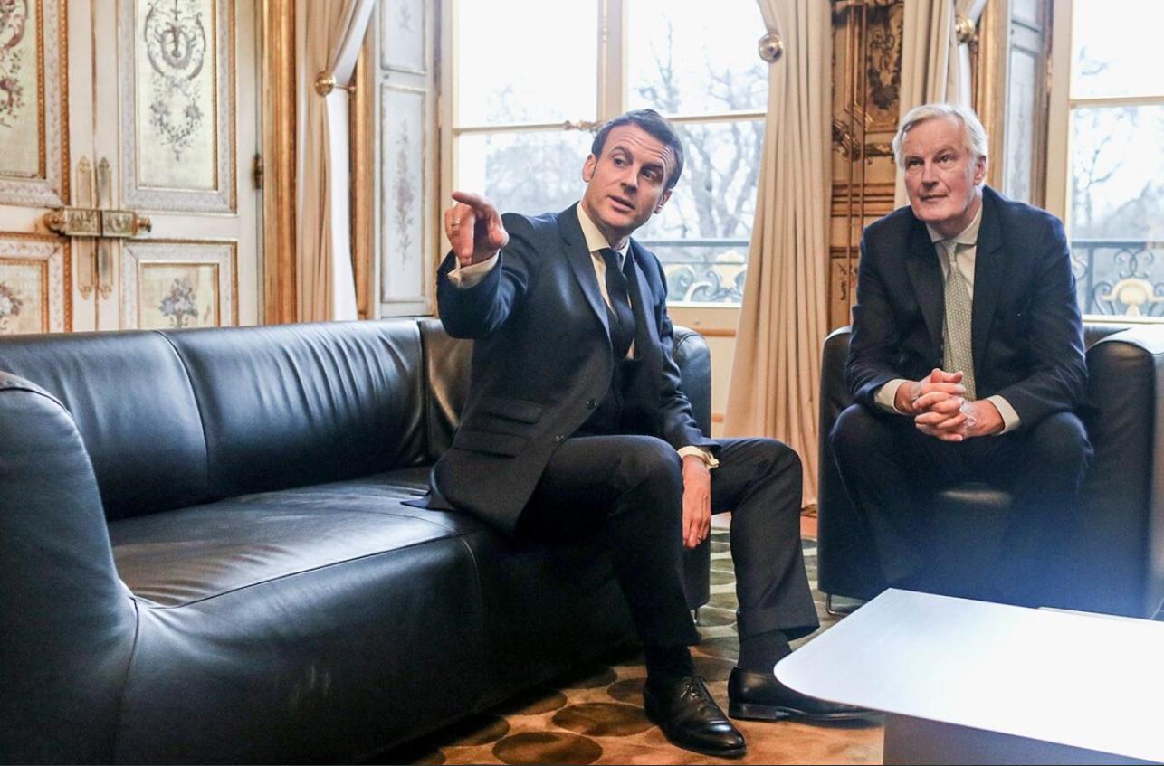 Michel Barnier et Emmanuel Macron assis sur des sièges en cuir. Emmanuel Macron pointe du doigt vers un objet hors champ.