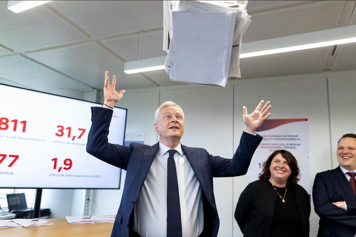 Bruno Le Maire, les bras levés, jette un paquet de documents format A4 en l'air devant deux personnes tout sourire et un en arrière-plan un écran présentant des chiffres.