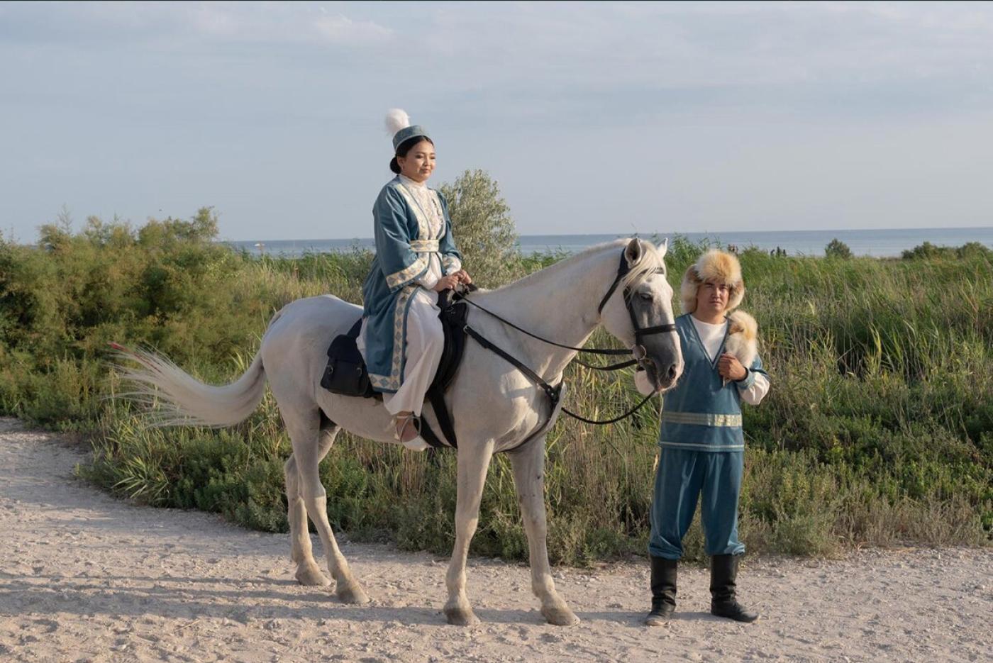 Une femme en tenue traditionnelle kazhak sur un cheval dont un homme tiens la bride sur un chemin de sable